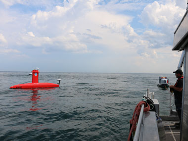 A red unmanned surface vessel on the ocean with a person controlling it from a nearby ship.