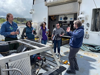 Four people listening to another person talk on the back deck of a ship