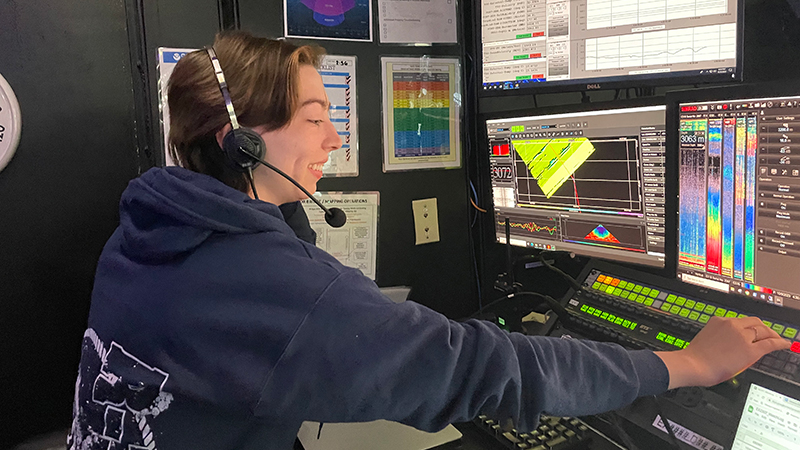 Person wearing a headset working at a control panel with multiple screens displaying graphs and charts.