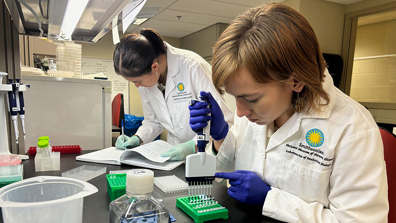 Two women in a lab using pipettes and taking notes.