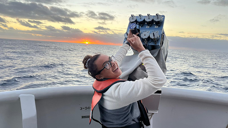 Person smiling on a boat holding a scientific instrument with a sunset over the ocean in the background.